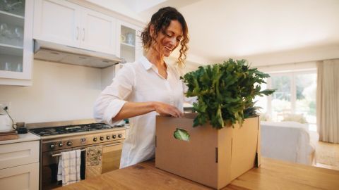 Eine Frau öffnet eine Kochbox in ihrer modernen Küche, um frisches Gemüse hervorzuzeigen und schnell zu kochen.