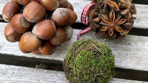 Drei Weihnachtskugeln aus Moos und Haselnüssen hängen auf einem Holztisch und verleihen festliche Atmosphäre.