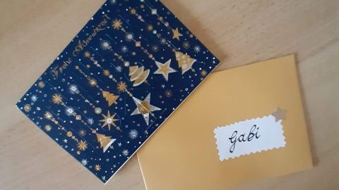 Ein satiniert gelber Briefumschlag mit dem Namen „Gabi“ liegt neben einer festlichen Karte mit goldenen Motiven.