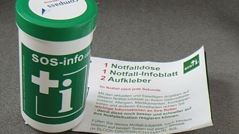 Eine grüne Notfalldose steht neben einem weißen Infoblatt, das wichtige Informationen für Notfallsituationen auflistet.
