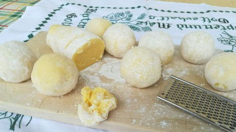 Runde, gelbe Klöße aus Kartoffelteig liegen auf einem Holzbrett, umgeben von Mehl und einer Reibe, bereit zum Kochen.
