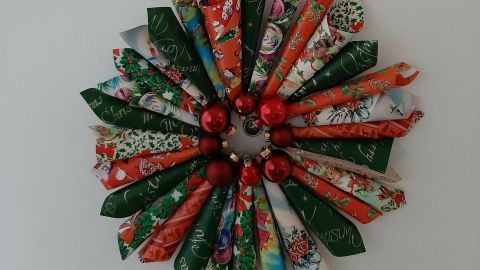 Ein bunter Weihnachtskranz aus Papierrollen und roten Kugeln hängt an einem roten Band an der Wand.