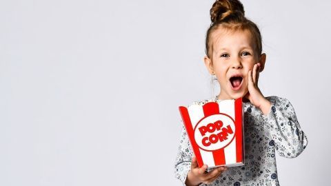Ein Mädchen mit Popcorn-Box überrascht mit offenem Mund, während sie in einem hellen Raum steht.