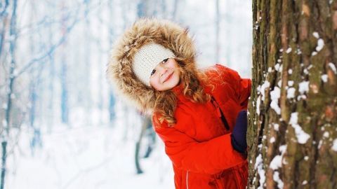 Ein Mädchen mit roter Jacke lehnt an einem Baum im verschneiten Winterwald und lächelt fröhlich.