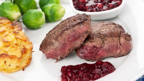 Zarte Rehrückenstücke liegen auf einem Teller, umgeben von bratanntem Kartoffelplätzchen, Rosenkohl und Preiselbeersoße.