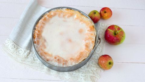 Ein gedeckter Apfelkuchen mit glattem Rum-Zuckerguss liegt in einer runden Form auf einem eleganten Tuch. Apfelhälften ergänzen die Szenerie.