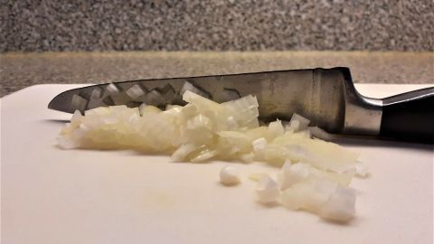 Ein scharfes Messer liegt auf einem Schneidebrett, umgeben von fein gehackten Zwiebelstücken.