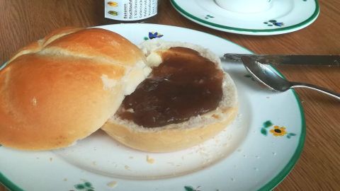 Ein Brötchen mit Bananenmarmelade liegt auf einem blumengeschmückten Teller, daneben ein Löffel und eine Tasse.