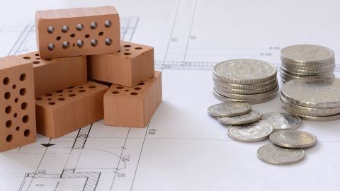 Ein Stapel von Bauziegeln steht neben einem Haufen Geldmünzen auf einem Bauplan, der das Hausbauprojekt darstellt.