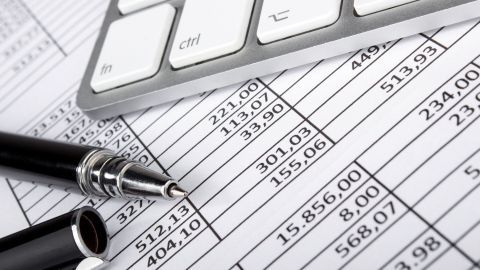 Eine schwarze Kugelschreibermine steht auf einem Finanzblatt, während eine Tastatur im Hintergrund bereitliegt, um Einnahmen zu verwalten.