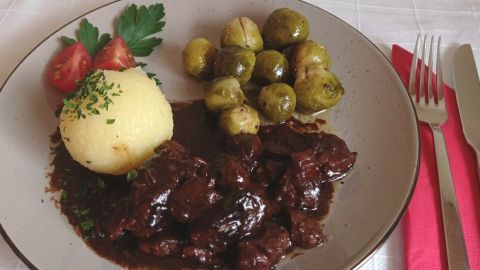 Ein Teller mit Schweinepfeffer, serviert mit Kartoffelknödel, Rosenkohl und Tomaten, auf einem weißen Tisch.