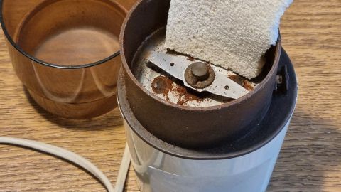 Eine Kaffeemühle steht auf einem Tisch, während ein Stück hartes Brot zur Reinigung im Mahlwerk positioniert ist.