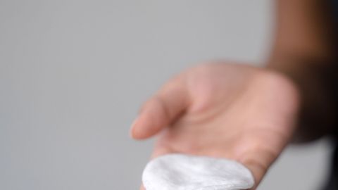 Eine Hand hält ein saugfähiges Wattepad, bereit zur Anwendung auf Pickeln, vor einem neutralen Hintergrund.