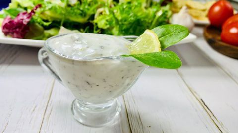 Ein Joghurtdressing in einer Glassoße mit einem Blatt Spinat und einer Zitronenscheibe steht vor einem frischen Salat.