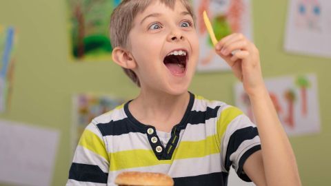 Ein junger Junge hält freudig eine Pommes frites in der Hand, während er vor einem Hamburger und Pommes sitzt.