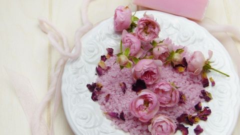 Auf einem weißen Teller liegt rosa Badesalz umgeben von frischen, rosa Rosenblüten und einer pinken Seife.