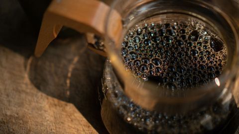 Ein Glas kalter Kaffee mit Bläschen steht auf einem Holztisch und reflektiert das Licht in der Umgebung.