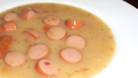 Ein cremiger Eintopf aus pürierter Brühe mit Scheiben von Würstchen schwimmt in einer weißen Schüssel.