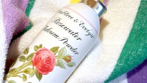 Verwende Rosenwasser-Talkumpuder, um Schweißfüße zu bekämpfen. Streue etwas auf deine Füße und in die Schuhe, um Feuchtigkeit und Gerüche zu reduzieren. Das Bild zeigt eine Dose Puder auf einem farbigen Handtuch als frische Lösung. Eine Dose Crabtree & Evelyn Rosewater Talcum Powder liegt auf einem bunten Handtuch. Die Dose zeigt eine Rose.
