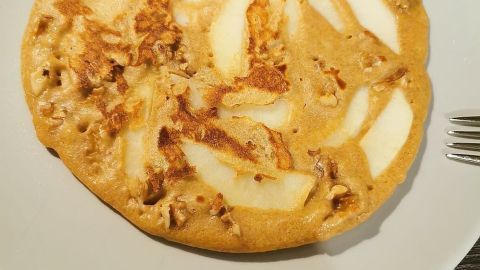 Ein gebratener Pfannkuchen aus Dinkelmehl, garniert mit Apfelstücken und Walnüssen, auf einem weißen Teller.
