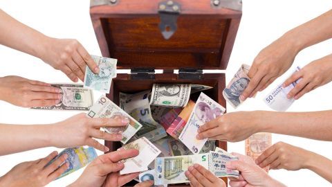 Eine Gruppe von Händen legt Geldscheine in eine offene Schatzkiste, die bereits mit verschiedenen Währungen gefüllt ist.