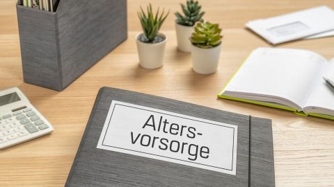 Ein graues Ordnersystem mit dem Titel "Altersvorsorge" liegt auf einem Holztisch, umgeben von Pflanzen und Bürobedarf.