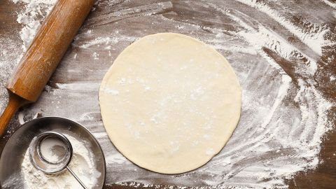 Ein runder Pizzateig liegt ausgerollt auf einer bemehlten Holzfläche neben einem Nudelholz und einem Sieb.