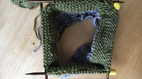 Eine gestrickte quadratische Fläche aus grünem und grauem Garn liegt auf einem Holztisch, umgeben von Wolle und Nadelspiel.