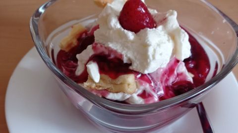 Eine Schüssel mit Quarkspeise wird mit roter Grütze, Sahne und einer Waffel dekoriert, auf einem Holzuntergrund präsentiert.