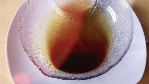 Eine klare Schüssel mit dunklem Kürbiskernöl steht auf einem weißen Teller, umgeben von gelben Unterlagen.