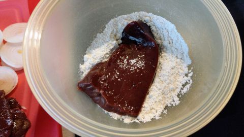 In einer Schüssel liegt ein Stück Fleisch in Mehl, während nebenan geschnittene Zwiebeln bereitstehen.