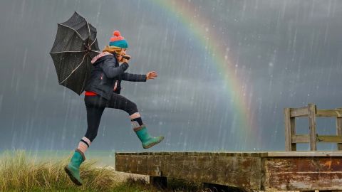 Eine Person springt lächelnd mit einem Regenschirm über eine Holzplanke, während es regnet und ein Regenbogen zu sehen ist.