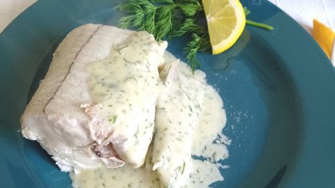 Gedünsteter Schellfisch liegt auf einem Teller, überzogen mit Kräuter-Béchamel und begleitet von einer Zitronenscheibe.