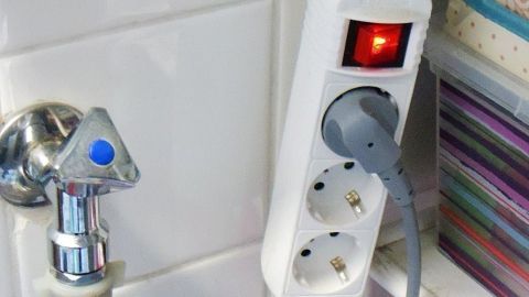 Ein weißer Verlängerungskabel mit einem angeschlossenen Stecker leuchtet rot, während es neben einem Wasserhahn in einem Badezimmer steht.