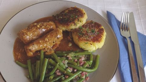 Saftige Involtini vom Schweinerücken liegen auf dem Teller, umgeben von Kartoffelplätzchen und grünen Bohnen.