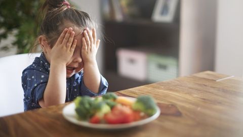 Ein Kind hält sich die Augen zu und zeigt eine abgeneigte Reaktion auf einen Teller mit Gemüse und Obst.
