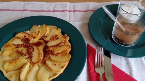 Ein Apfelpfannkuchen liegt auf einem grünen Teller, umgeben von Besteck und Zucker im Hintergrund.