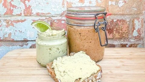 Ein rustikaler Brotaufstrich liegt auf einem Holzbrett, umgeben von zwei Gläsern mit veganen Aufstrichen vor einer Backsteinwand.