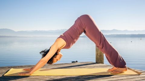 Eine Frau führt im herabschauenden Hund Yoga am Ufer eines ruhigen Sees bei Sonnenlicht aus, umgeben von Bergen.
