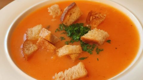 Eine cremige Karotten-Kartoffelsuppe steht in einer weißen Schüssel, garniert mit Croûtons und frischem Grün, auf einem Holztisch.