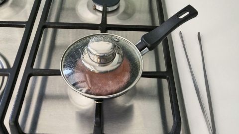 Ein Burger-Patty wird in einer kleinen Pfanne mit Deckel auf einem Gasherd gebraten, umgeben von Edelstahloberflächen.