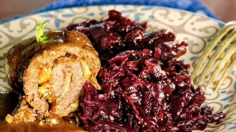 Für besonders zarte Rouladen werden diese flach und breit geklopft, mit Öl eingerieben, übereinandergelegt, in Frischhaltefolie gewickelt und im Kühlschrank eine Nacht gelagert. Zarte Rouladen liegen neben einem kräftigen Rotkohl auf einem dekorativen Teller, umgeben von einer rustikalen Tischumgebung.