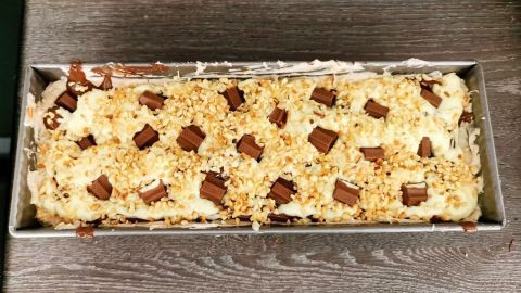 Ein üppiger Kinderriegel-Kuchen liegt in einer Backform, bedeckt mit Schokoladenstücken und gehackten Nüssen.