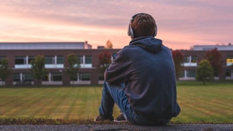 Ein Junge sitzt mit Kopfhörern auf einer Mauer und schaut auf eine Wiese vor einem Schulgebäude bei Sonnenuntergang.