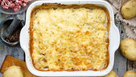 Ein goldbrauner Püree-Sauerkrautauflauf liegt heiß in einer weißen Auflaufform auf einem rustikalen Holztisch, umgeben von Zutaten.