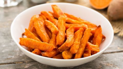 Süßkartoffel-Pommes liegen knusprig in einer weißen Schüssel auf einem rustikalen Holztisch, daneben ein Stück Süßkartoffel.