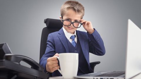 Ein Junge sitzt in einem Bürostuhl und hebt eine Tasse, während er an einem Laptop arbeitet und konzentriert blickt.