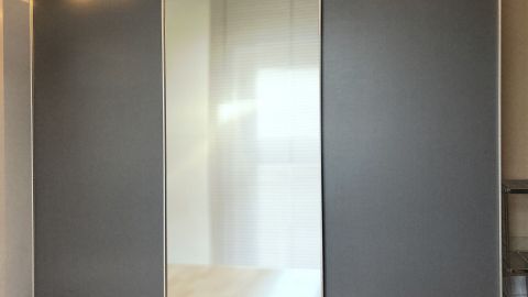 Ein großer, moderner Kleiderschrank mit einem Spiegel in der Mitte steht in einem hellen Raum mit Holzfußboden.