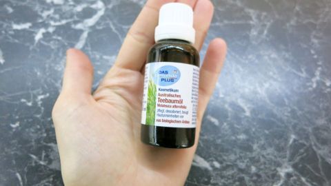 Eine Hand hält eine Flasche mit Teebaumöl, was auf eine mögliche Anwendung bei Schnupfen hinweist. Der Hintergrund ist grau marmoriert.