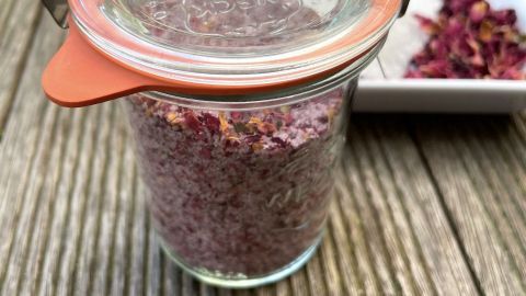 Ein Glas mit Rosensalz steht auf einem Holztisch, während getrocknete Rosenblätter daneben liegen.